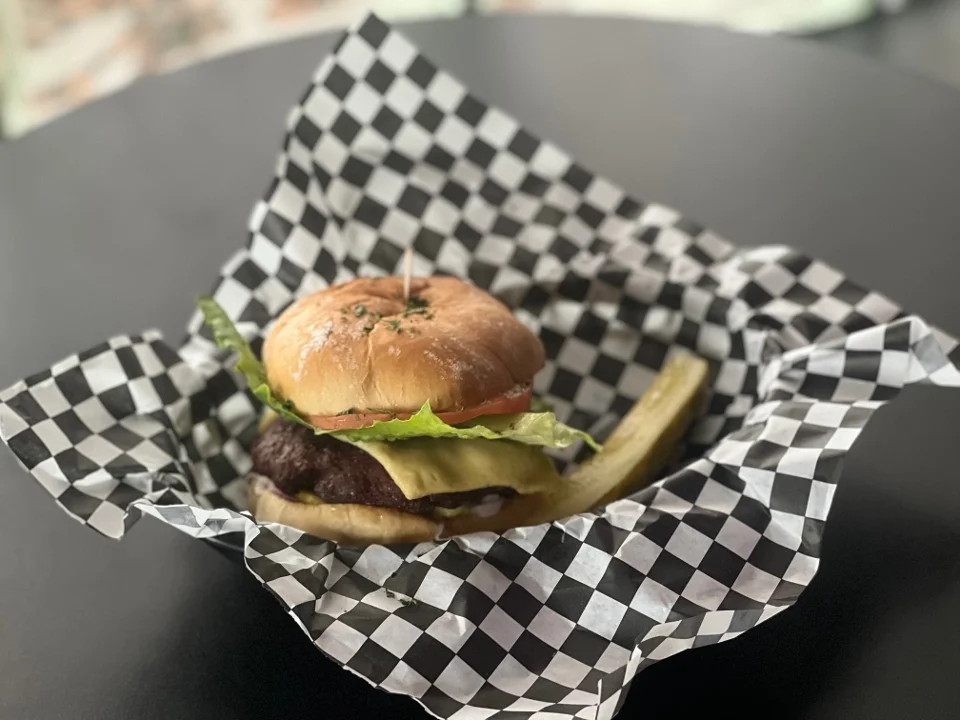 A juicy hamburger with lettuce, tomato, and cheese served in a checkered paper basket alongside a pickle.