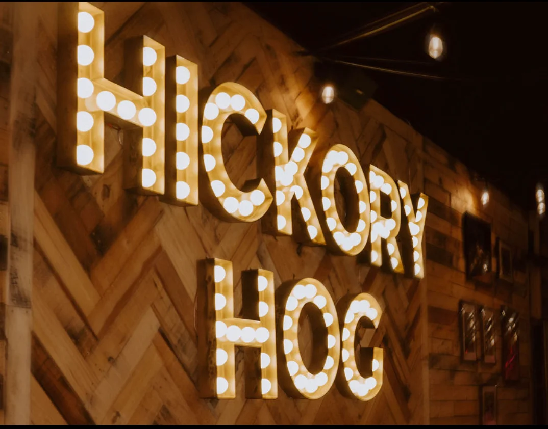 A rustic wooden wall featuring illuminated letters spelling 'HICKORY HOG'.