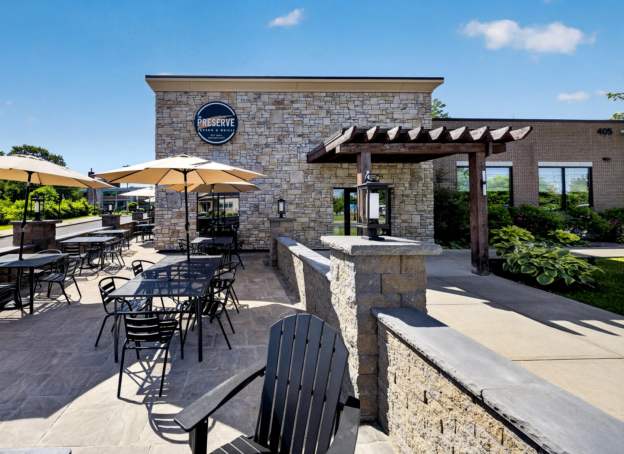 Sunny outdoor stone patio at The Preserve Tavern & Grille, featuring black metal dining tables with beige umbrellas, stone walls, a wooden pergola walkway, and lush green landscaping under a bright blue sky.
