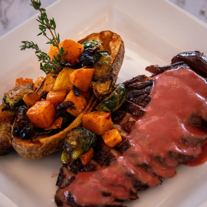 A gourmet dish featuring sliced steak with a sauce, accompanied by roasted vegetables and a baked potato half.