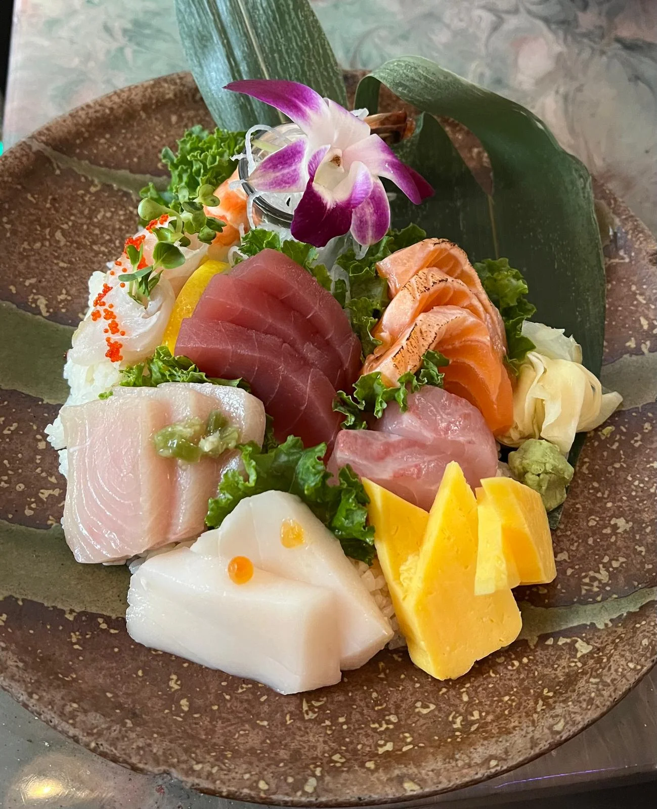 A colorful sashimi platter featuring various types of fish, including salmon and tuna, garnished with greens and an orchid flower.