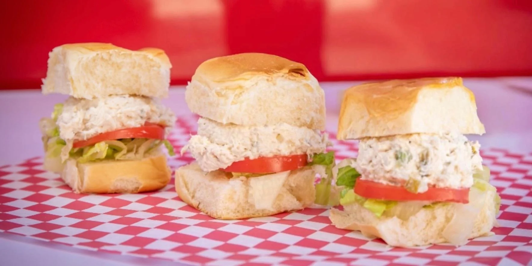 Three mini sandwiches made with soft bread, filled with chicken salad, lettuce, and tomato, placed on checkered red and white paper.