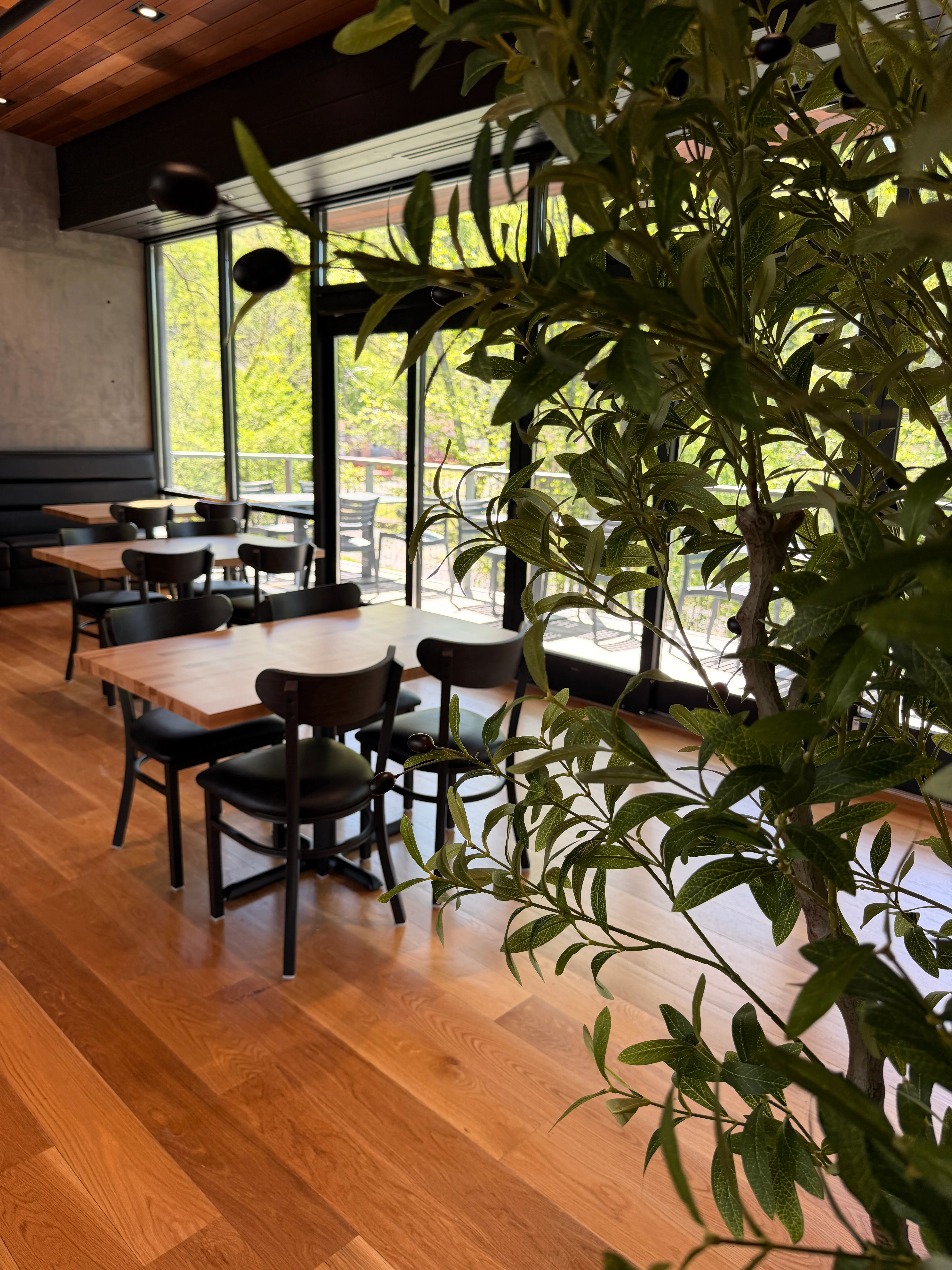 A spacious restaurant interior with several wooden tables and black chairs, brightened by large windows and decorated with wall art.