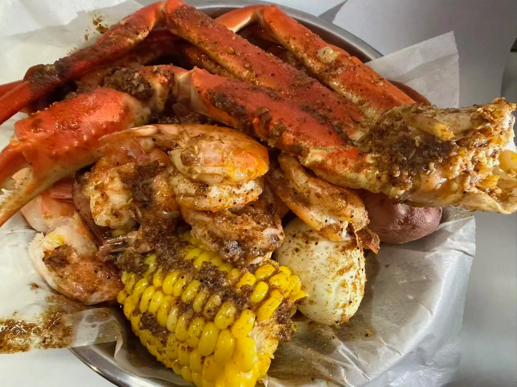 A bowl containing a flavorful mix of seafood including crab legs, shrimp, corn, and seasoned potatoes, served with a side of paper.