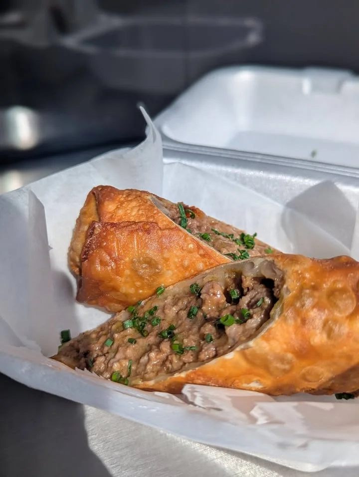 Two golden-brown meat-filled pastries served in a paper container, garnished with chopped green herbs.