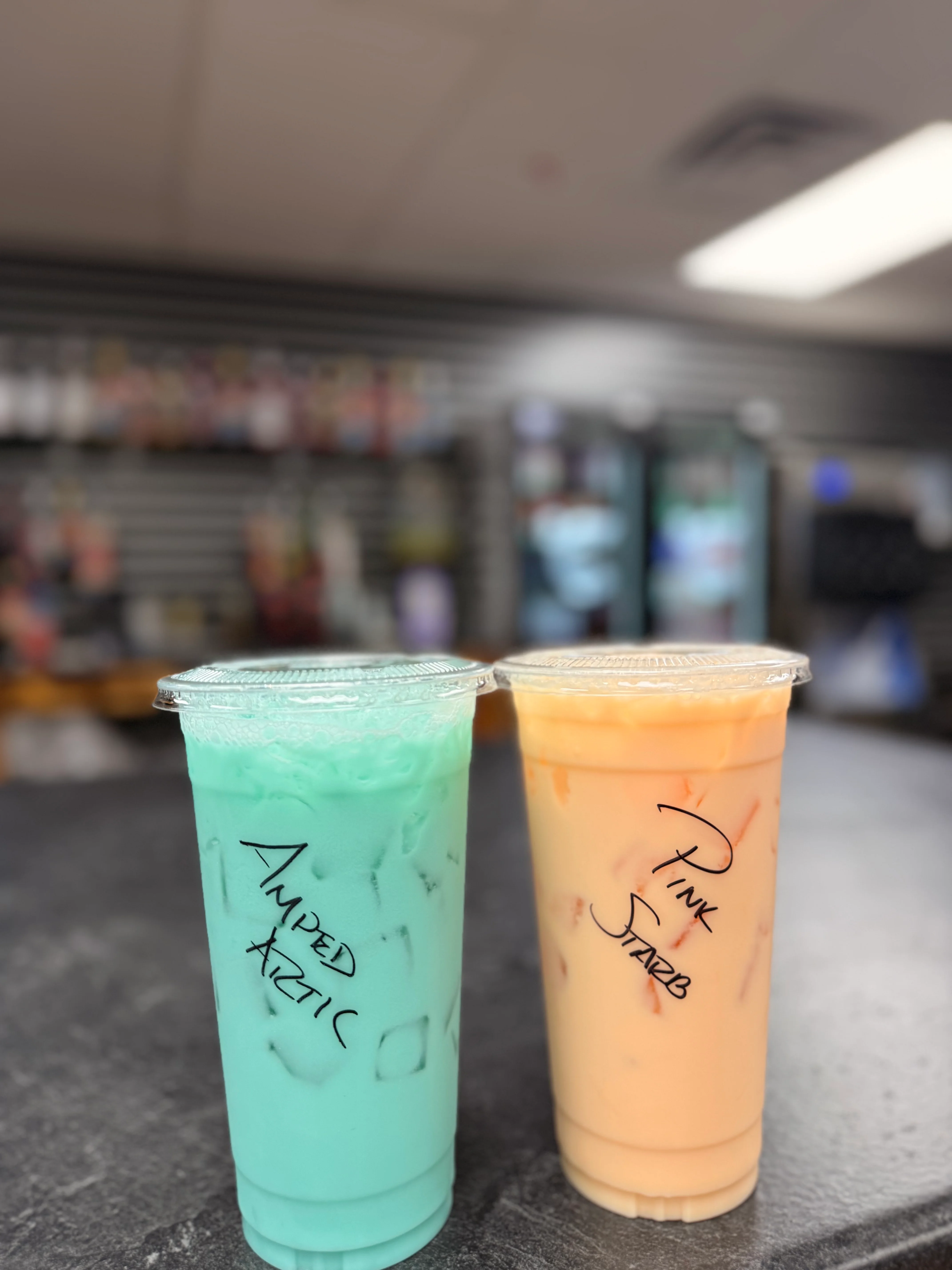 Two vibrant drinks in clear cups, showcasing bright colors and unique names, on a store countertop.