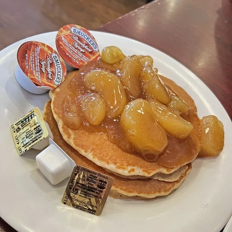 A stack of fluffy pancakes topped with glazed apples and syrup, served with packets of butter and breakfast syrup on a white plate.