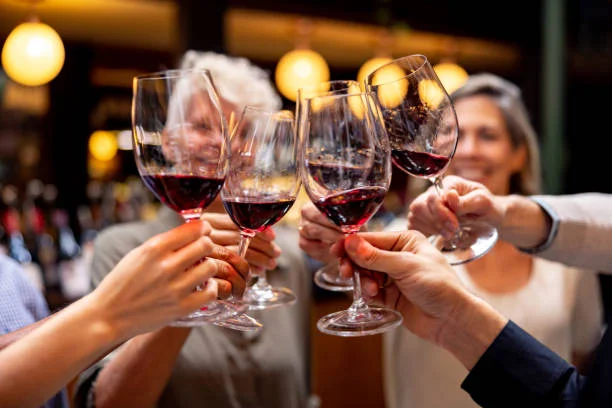 A group of people clinks glasses of red wine in a celebratory toast. The background is softly lit with warm, golden lights, creating a festive atmosphere.