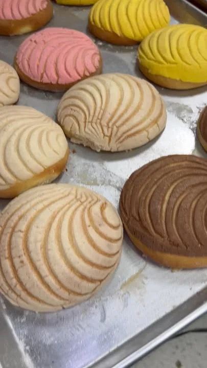 A tray filled with various colorful conchas, a type of sweet Mexican bread, featuring pink, yellow, and brown tops with a distinctive spiral pattern.