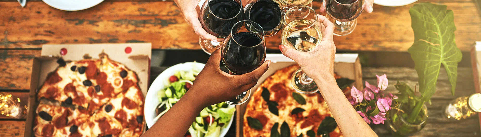 People toasting with glasses of wine over a table with pizza and salad on it.