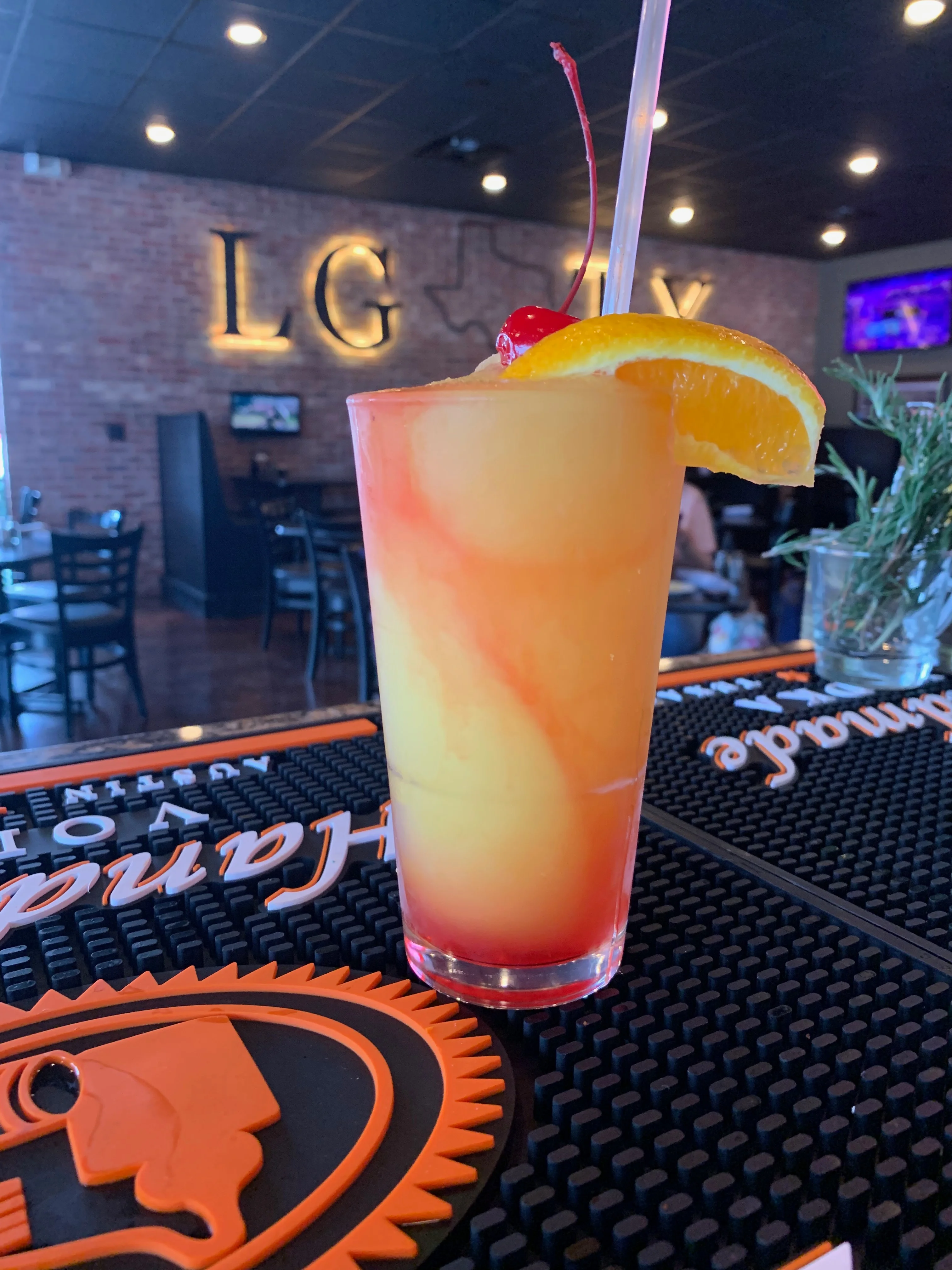 A colorful tropical cocktail with layers of orange and red, garnished with a cherry and an orange slice, served in a tall glass.