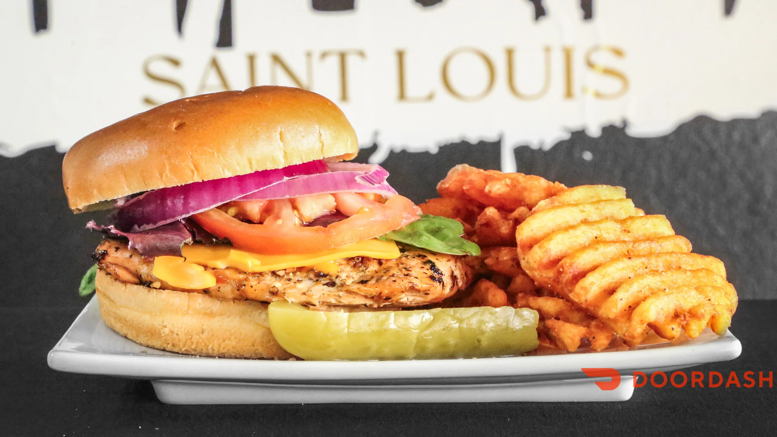 A close-up of a chicken burger topped with lettuce, tomato, onion, and cheese, served with waffle fries and a pickle on a plate.