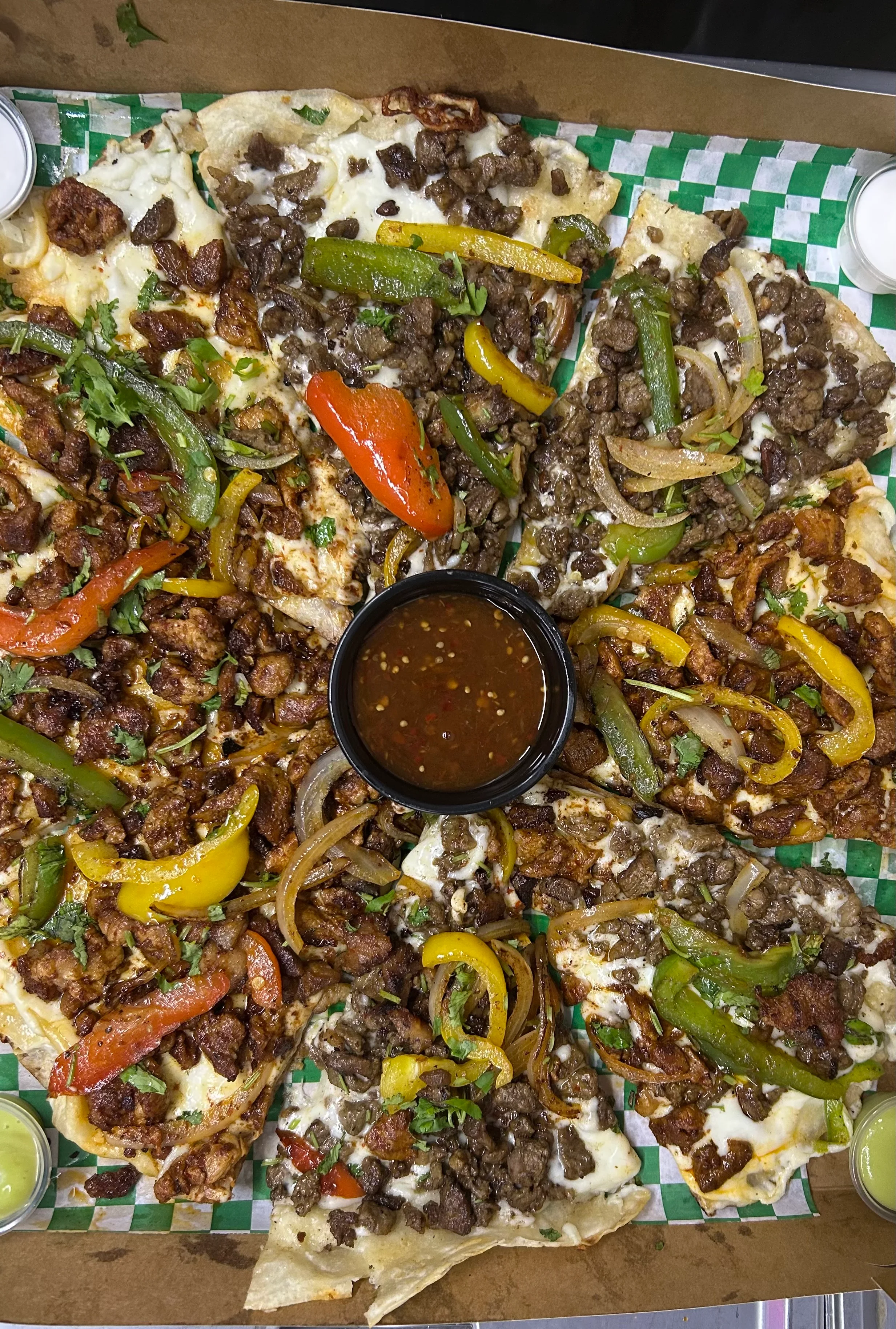 A tray of nachos topped with various meats, bell peppers, onions, and cheese. A small bowl of sauce is placed in the center.