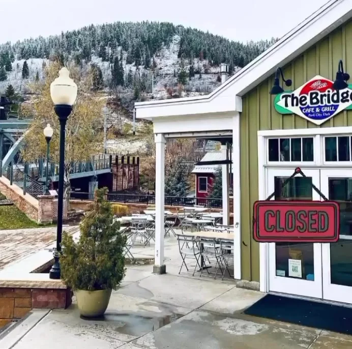 An outdoor view of a cafe with a closed sign, surrounded by tables and greenery. Snow-covered hills are seen in the background.