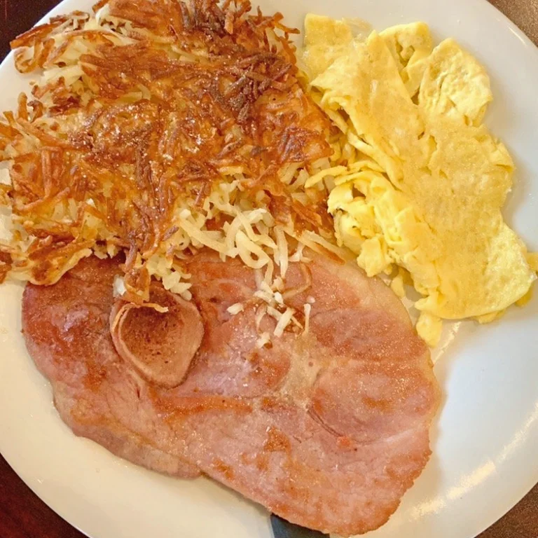 A thick ham steak with scrambled eggs and a side of crispy hashbrowns, presented on a white plate.