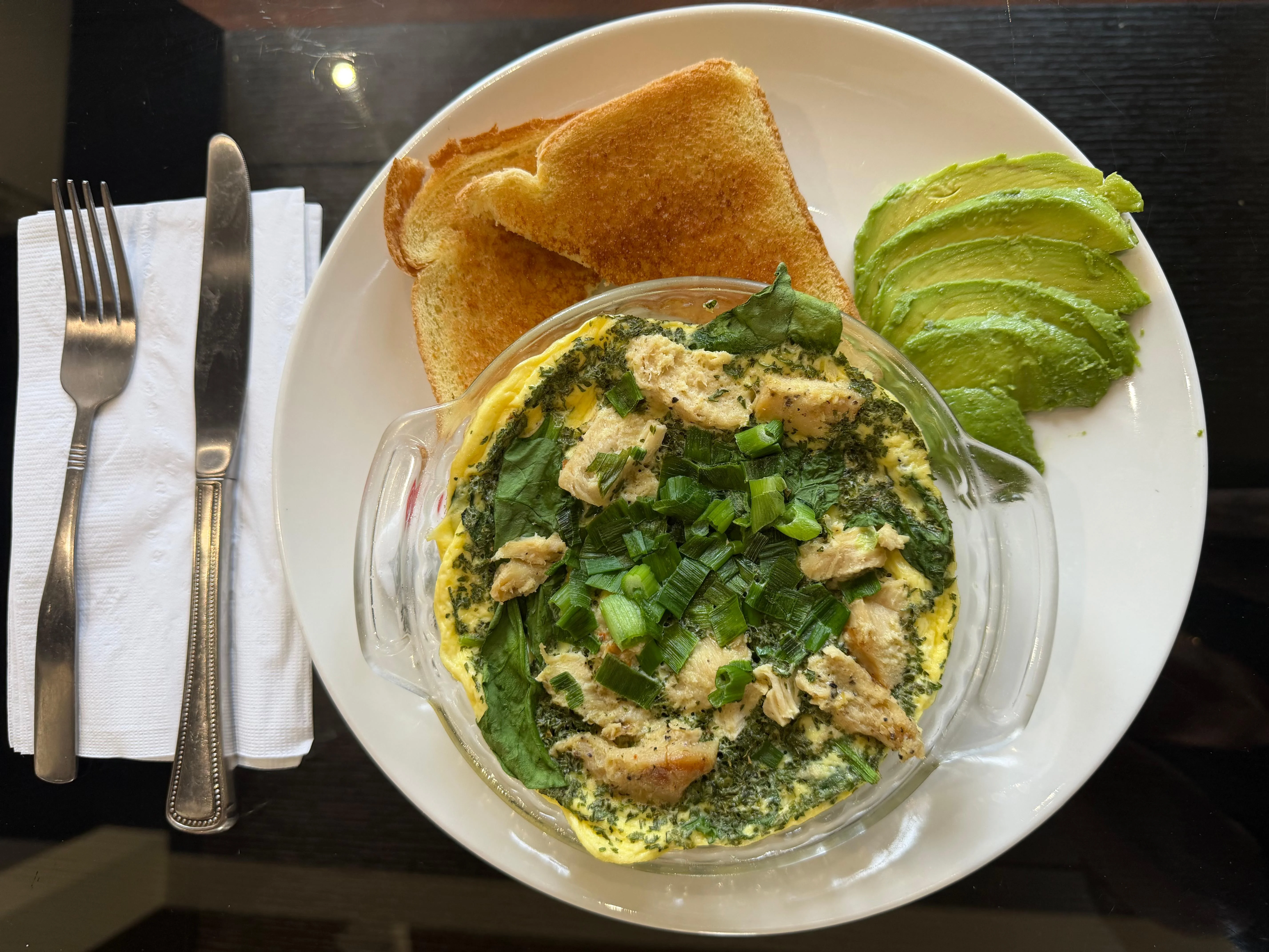 A serving of baked egg dish with spinach and chicken, topped with green onions, served alongside toast and sliced avocado.