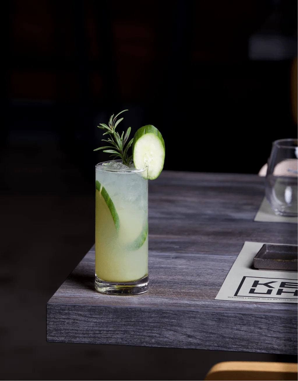 A refreshing highball cocktail served over ice, garnished with a cucumber slice and a sprig of fresh rosemary, resting on the corner of a dark wooden restaurant table.