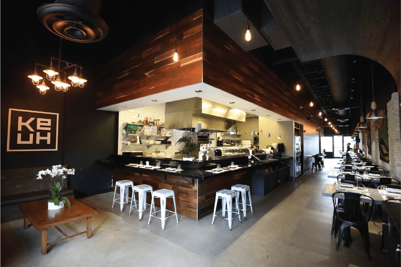Modern restaurant interior featuring a central wooden sushi bar with white industrial stools, black tables along a brick wall, and warm Edison bulb lighting.