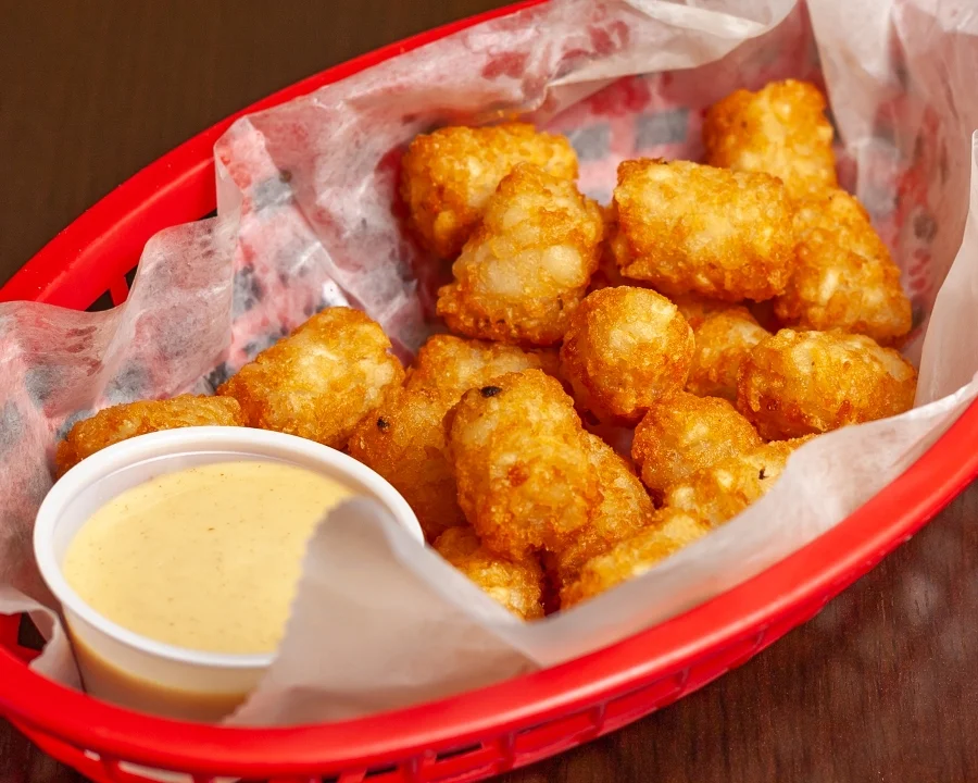 A red basket filled with golden tater tots, accompanied by a small cup of yellow sauce.