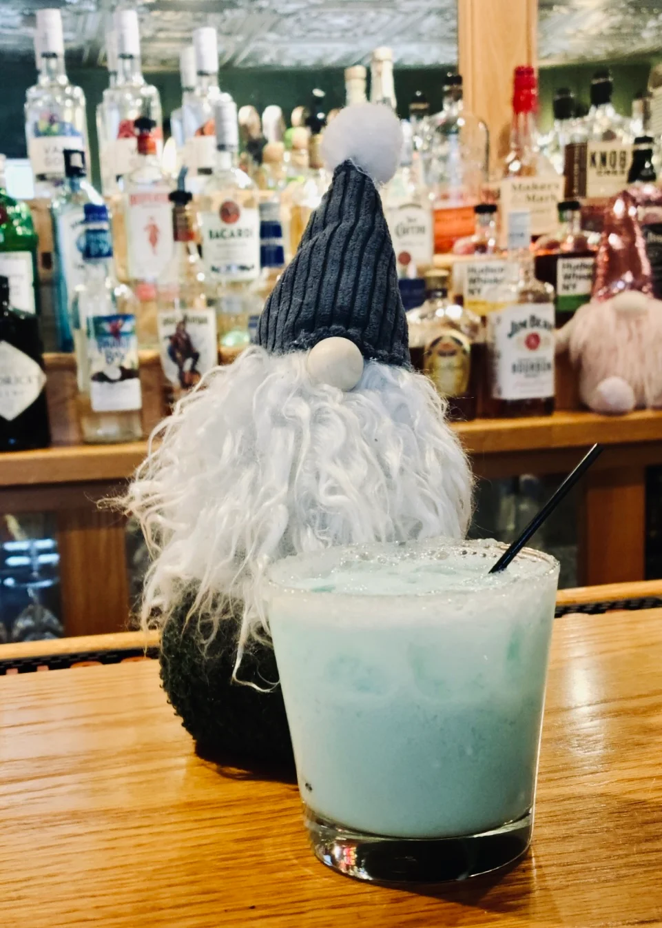 A whimsical gnome figure stands next to a vibrant blue cocktail with a straw. Behind, a well-stocked bar is visible, filled with various bottles.