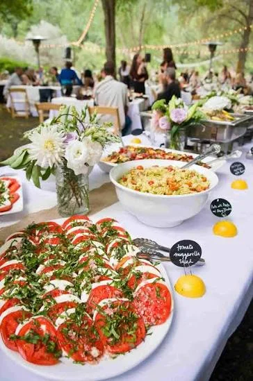 An outdoor buffet spread with fresh salads, featuring colorful ingredients and floral arrangements, set amid a lively gathering.