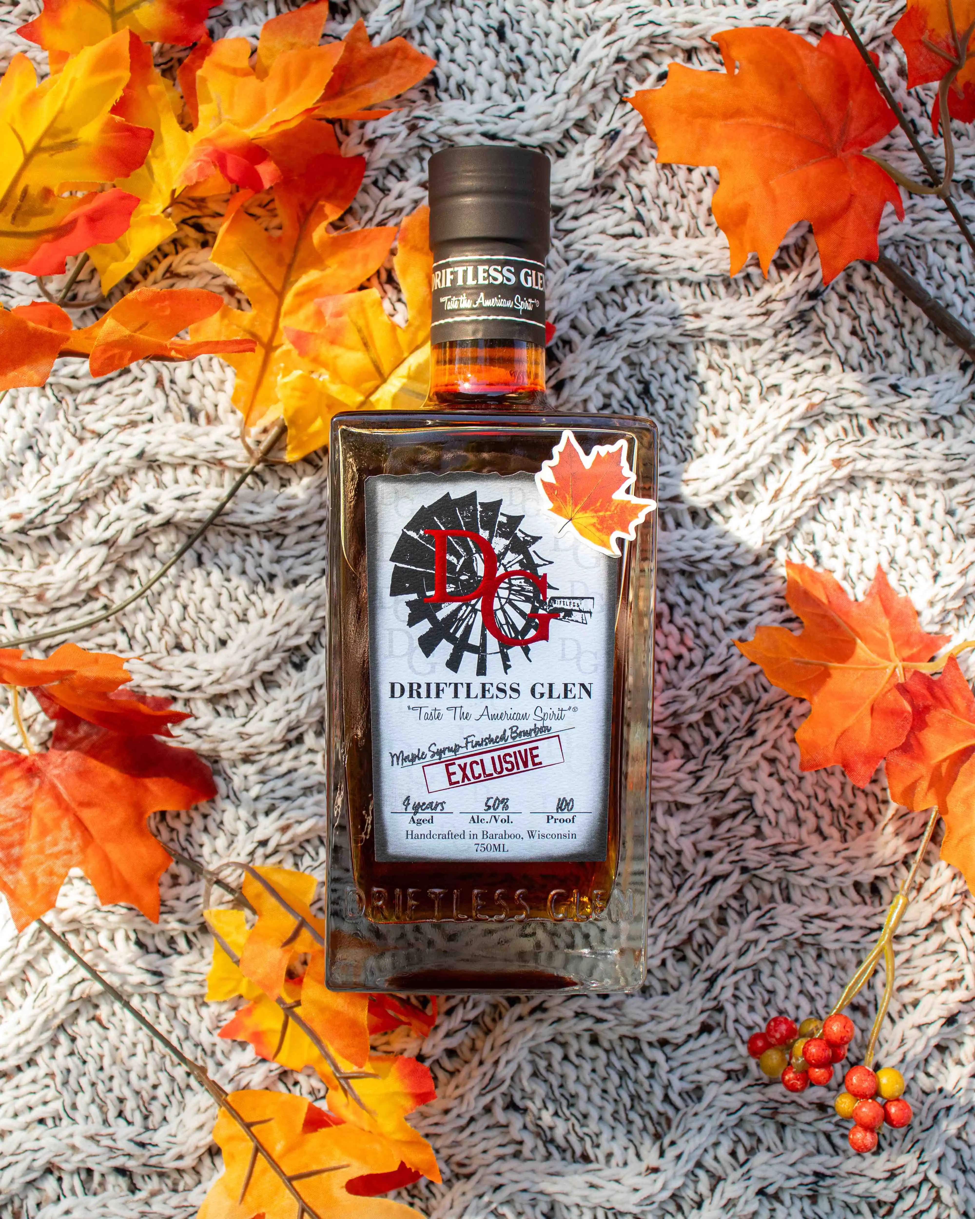 A bottle of Maple Syrup-Finished Bourbon sits on a cozy knitted blanket with orange, red, and yellow leaves strewn around.