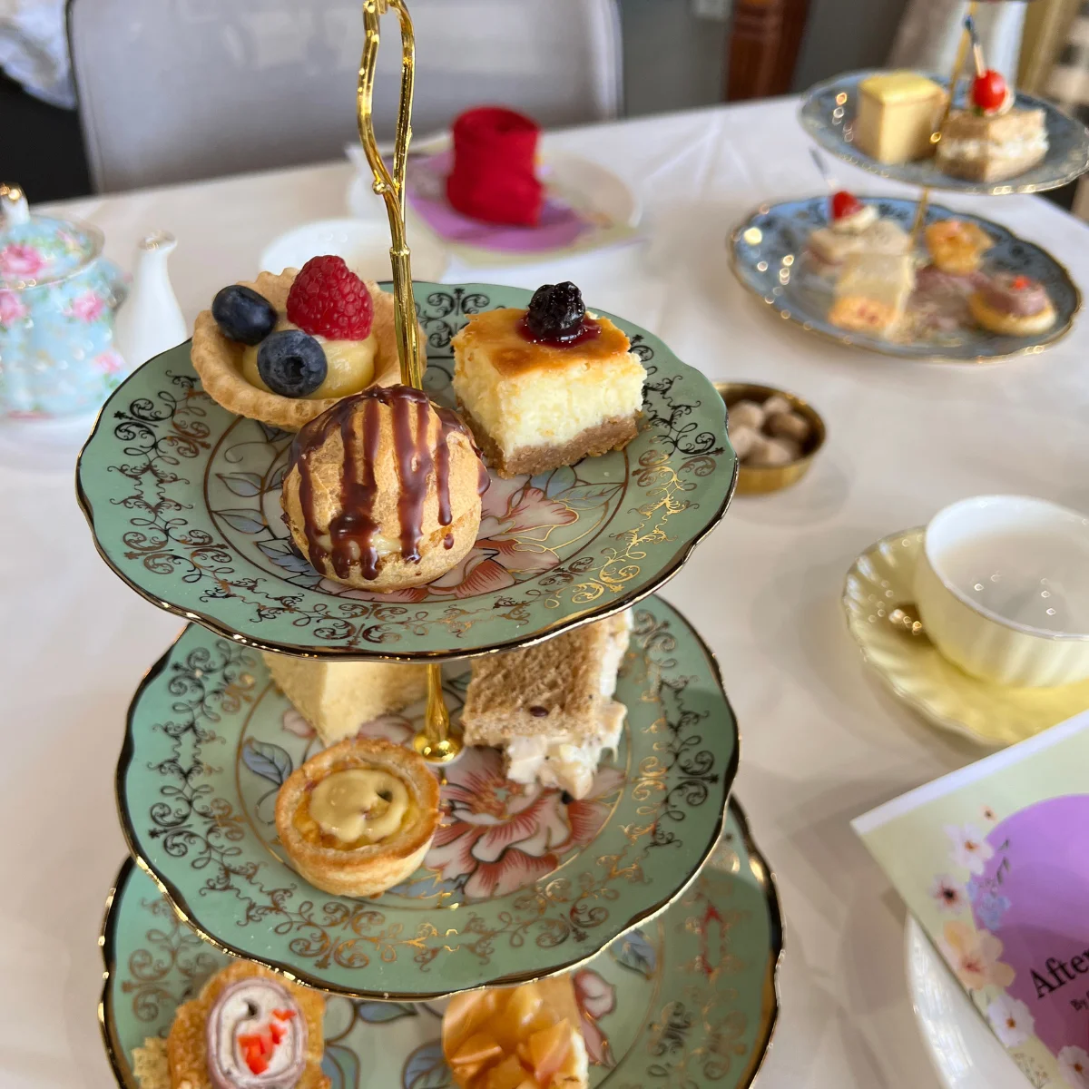 A three-tiered tray of assorted afternoon tea treats, including fruit tartlets, cheesecake, cream puffs, and finger sandwiches, served on elegant floral china with gold trim.
