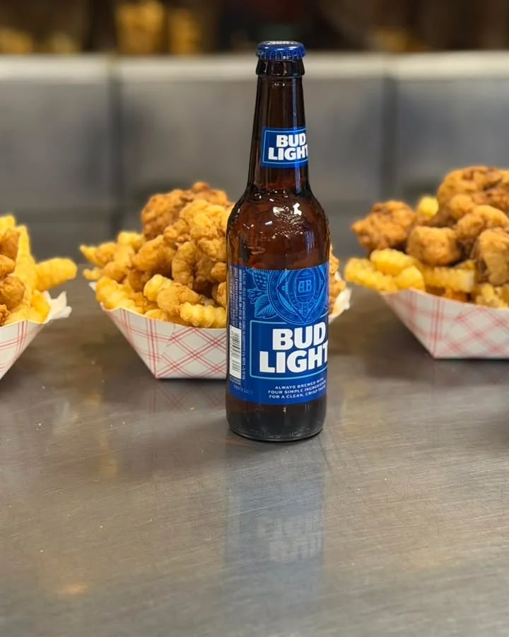 A bottle of beer placed in front of two servings of crinkle-cut fries and fried chicken. The food is served in paper trays.