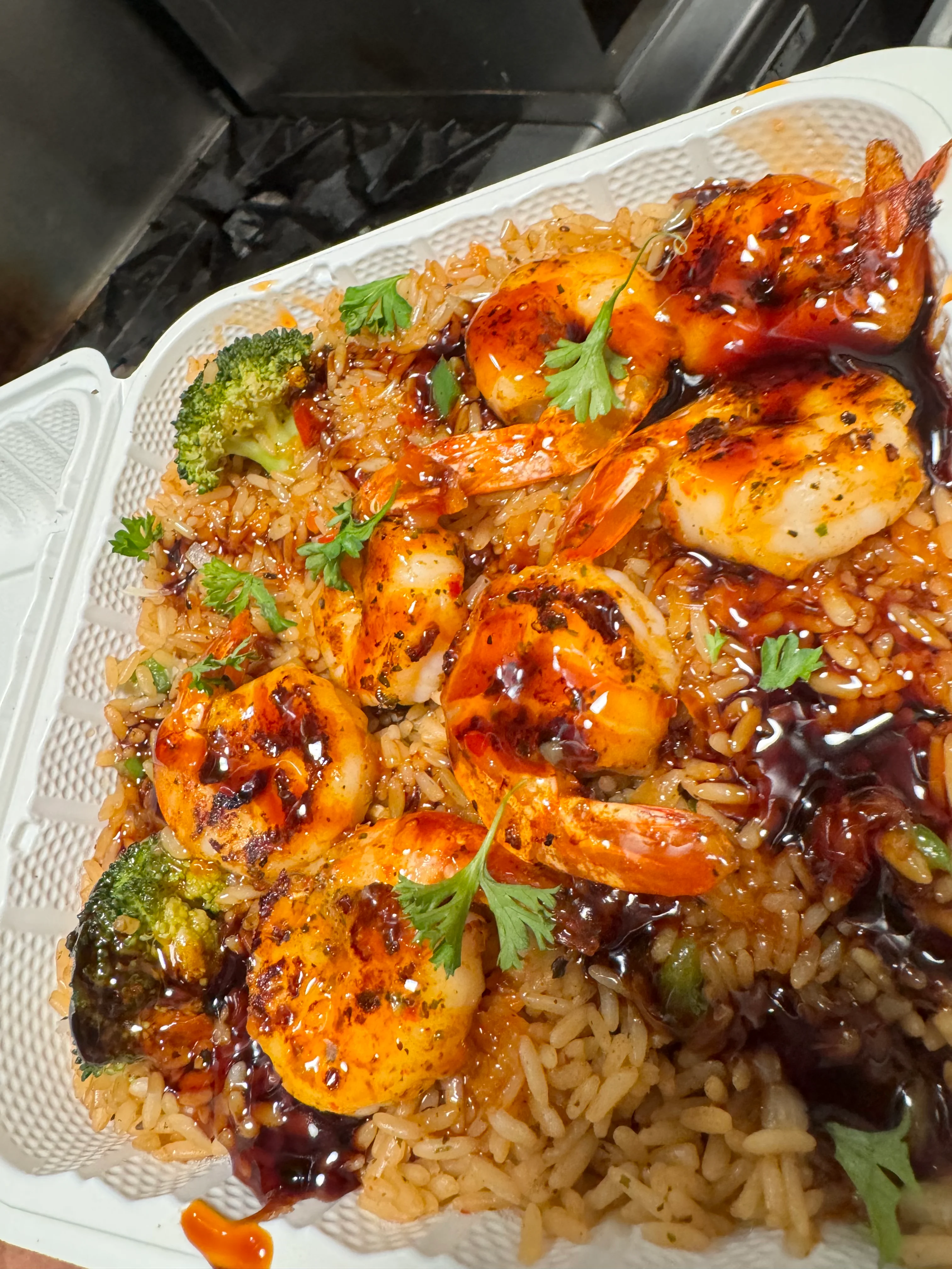 A delicious meal featuring grilled shrimp served over rice, garnished with broccoli and cilantro in a takeout container.