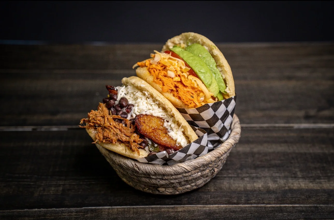 Three arepas filled with various ingredients, including pulled chicken, cheese, black beans, and avocado, served in a woven basket.