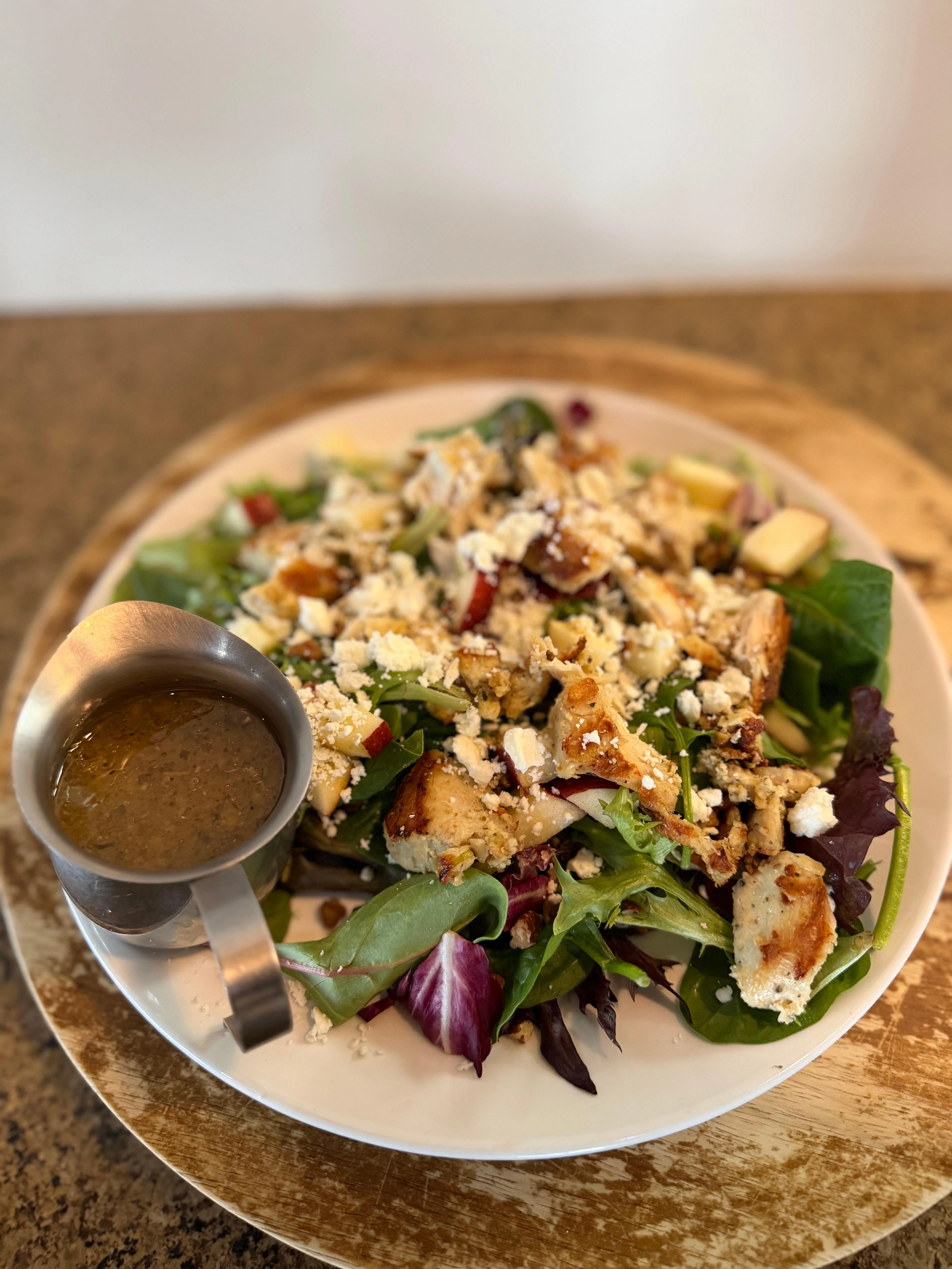 A colorful salad with assorted greens, diced apples, and croutons topped with crumbled cheese and served with dressing.