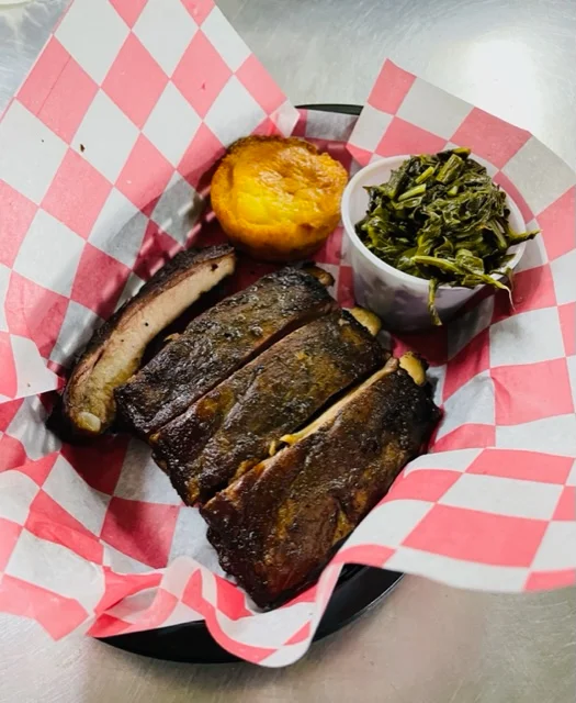 A plate featuring tender barbecue ribs alongside a cornbread muffin and a serving of greens, all arranged with a red and white checkered paper.