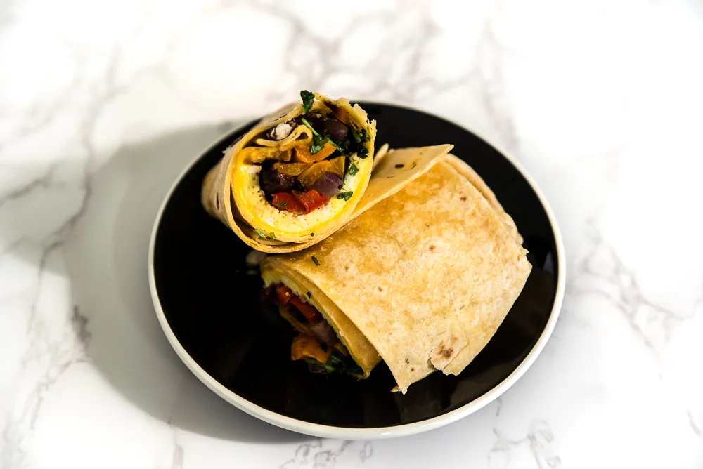 A wrap sandwich on a black plate, split to show colorful fillings like grilled vegetables, greens, and cheese, on a white marble background.