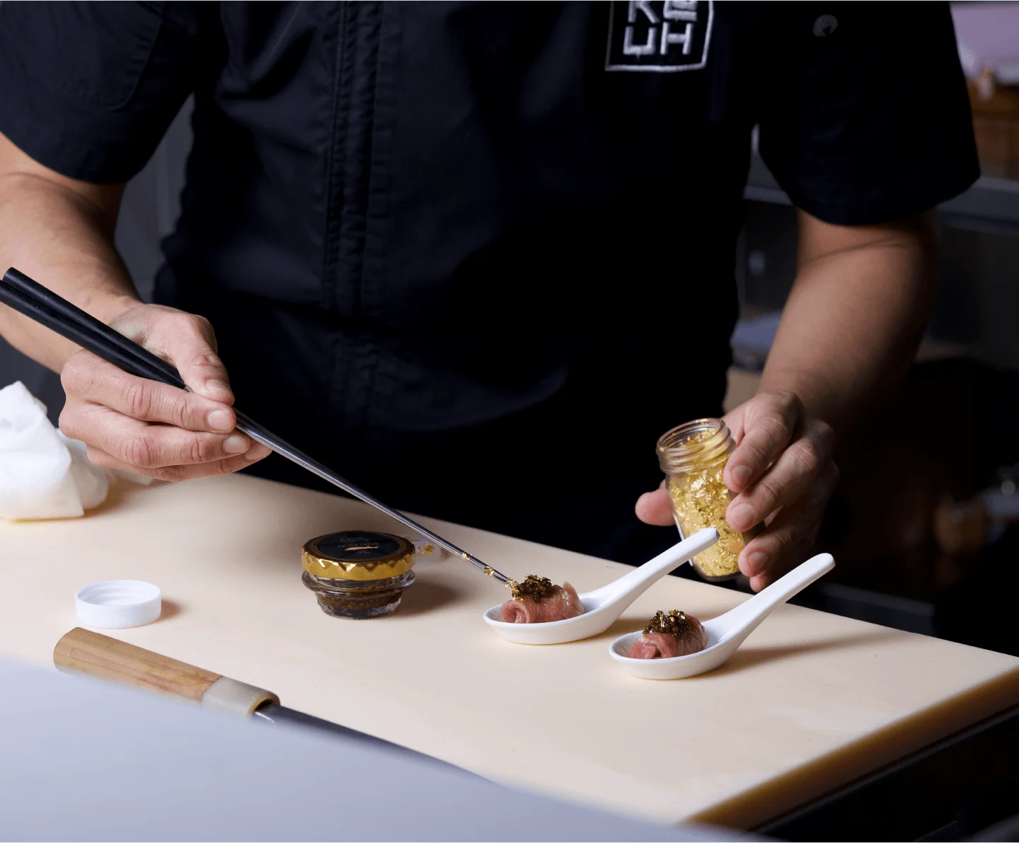 A chef wearing a branded Ke-uH uniform uses long metal chopsticks to delicately place a combination of caviar and edible gold flakes onto small, rolled appetizers resting in white ceramic spoons.