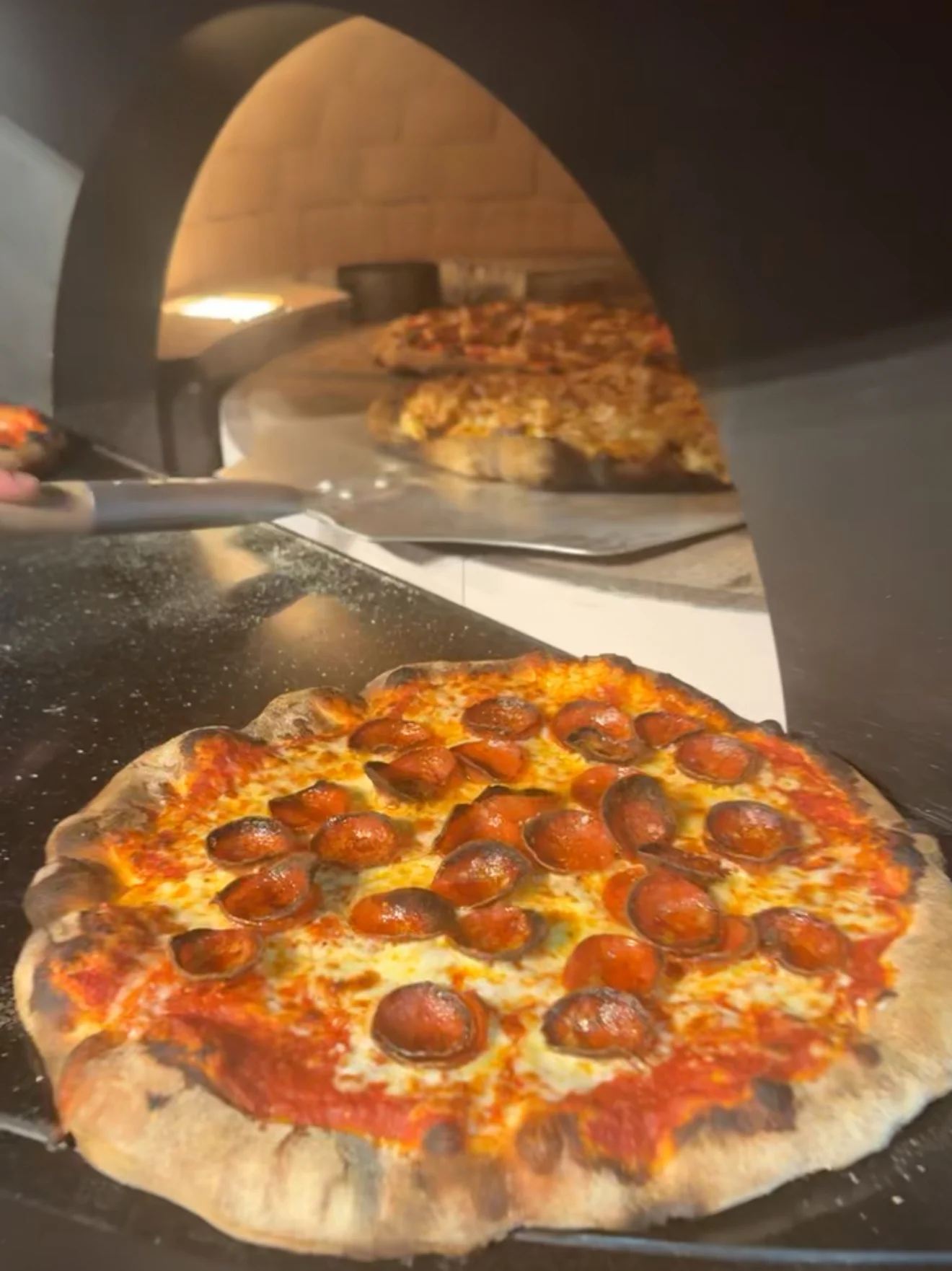 A freshly baked pepperoni pizza with a crispy crust, placed in a wood-fired oven. The pizza is topped with red sauce and melted cheese, showcasing the toppings distinctly.