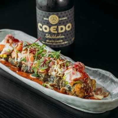 A platter of sushi rolls drizzled with sauce and garnished with microgreens, accompanied by a bottle of craft beer.