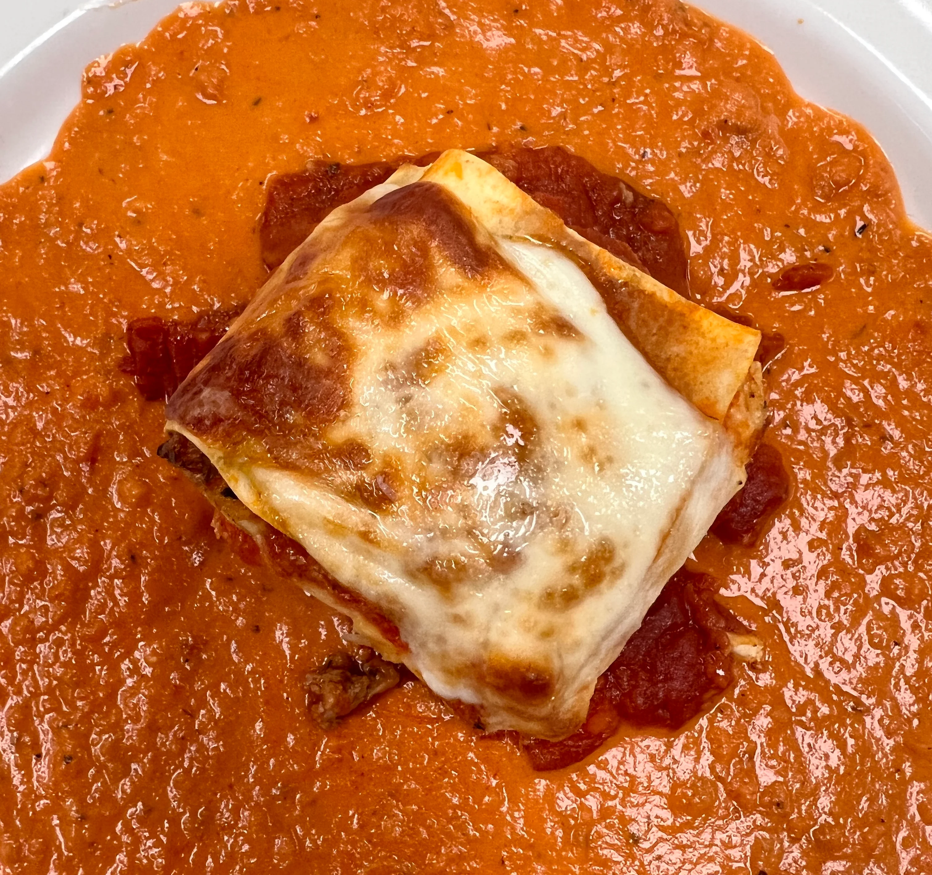 A square portion of baked lasagna with golden, melted mozzarella cheese on top, served in the center of a white plate surrounded by a generous pool of rich, creamy tomato sauce. The lasagna layers and hearty filling are visible on the sides beneath the cheese.