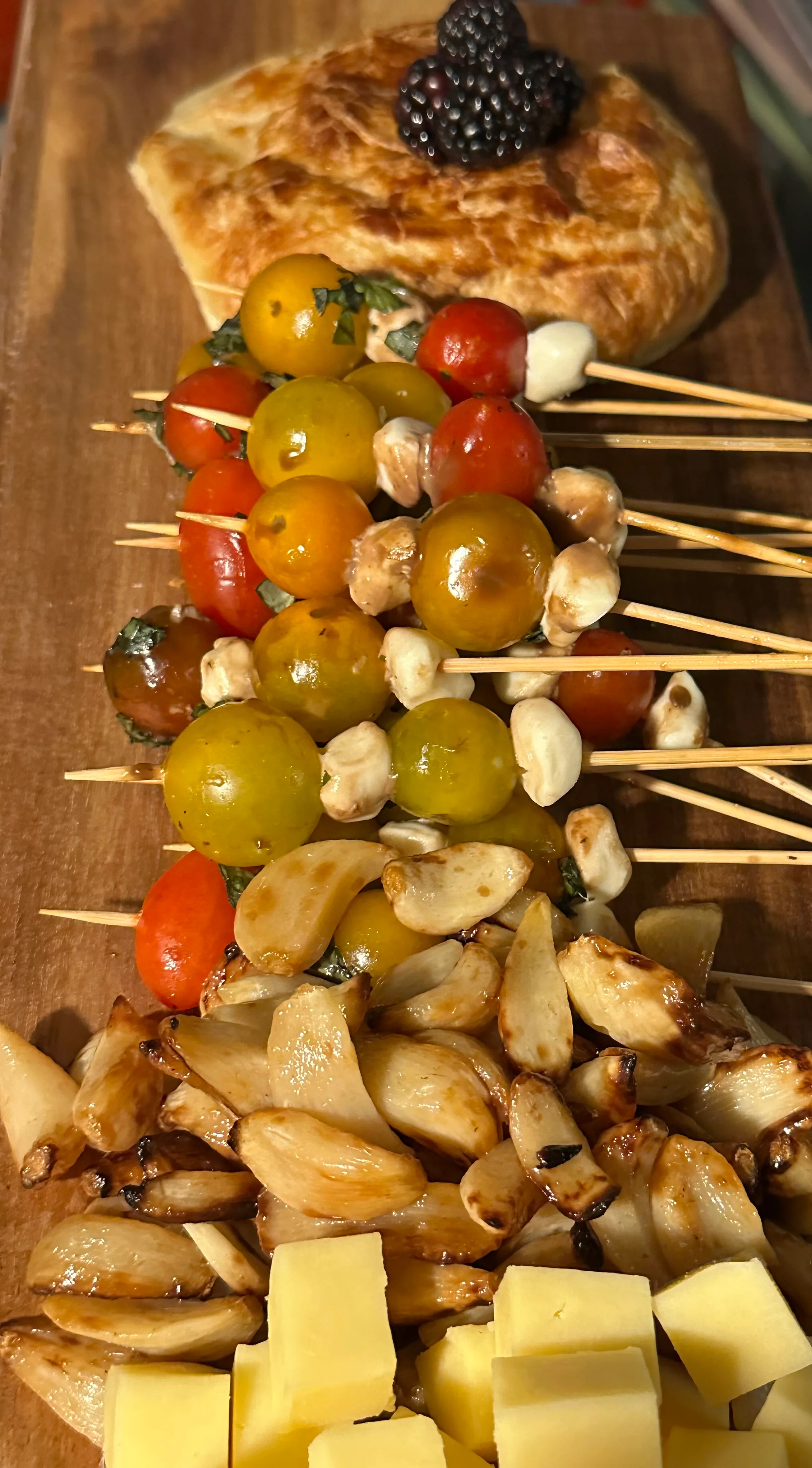 A wooden serving board featuring a variety of food items including skewers with cherry tomatoes and cheese, roasted garlic, cubes of cheese, and a pastry topped with a blackberry.
