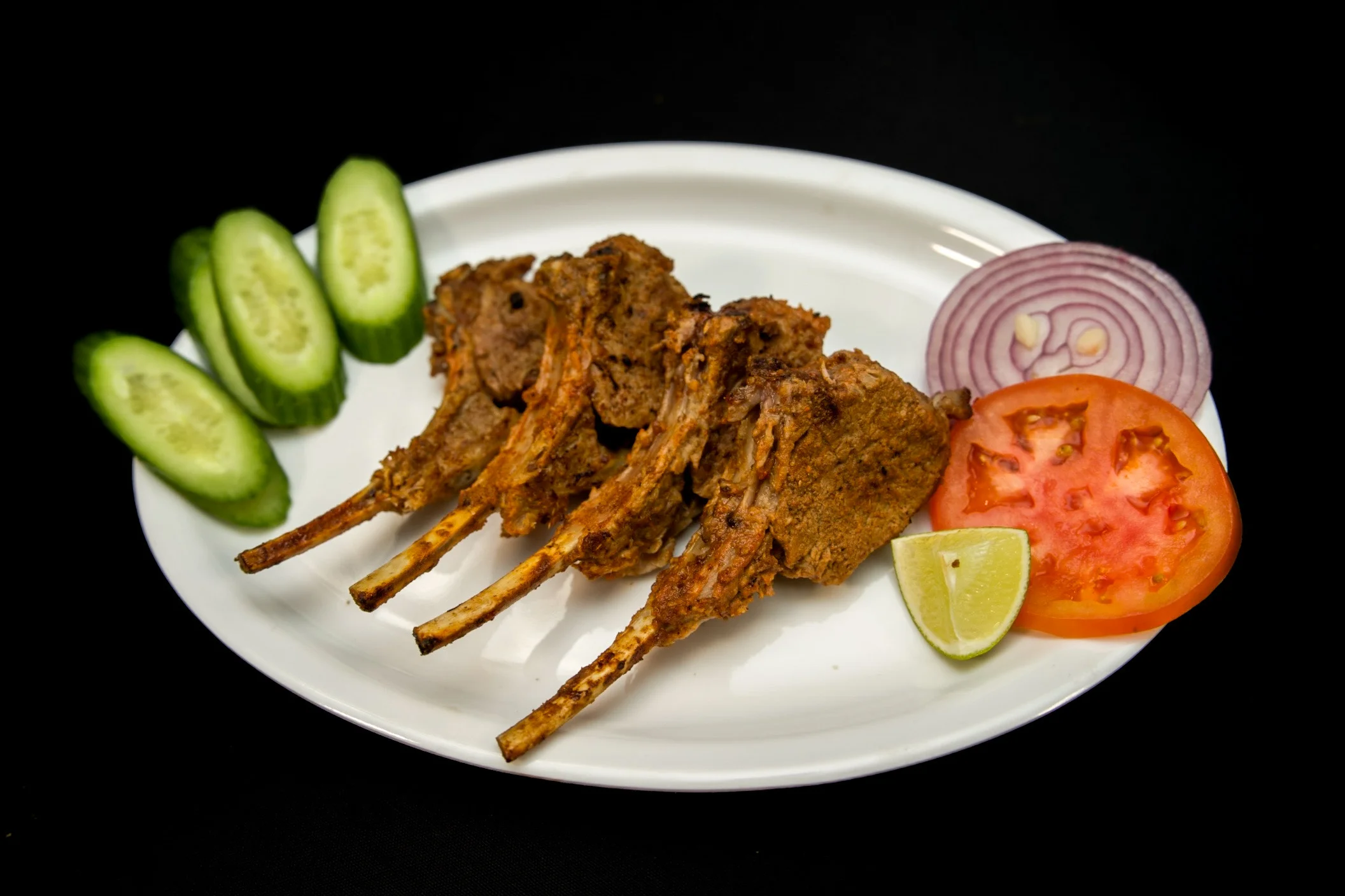 Grilled mutton chops served with sliced cucumber, tomato, red onion, and a lime wedge at Karachi BBQ Tonight.