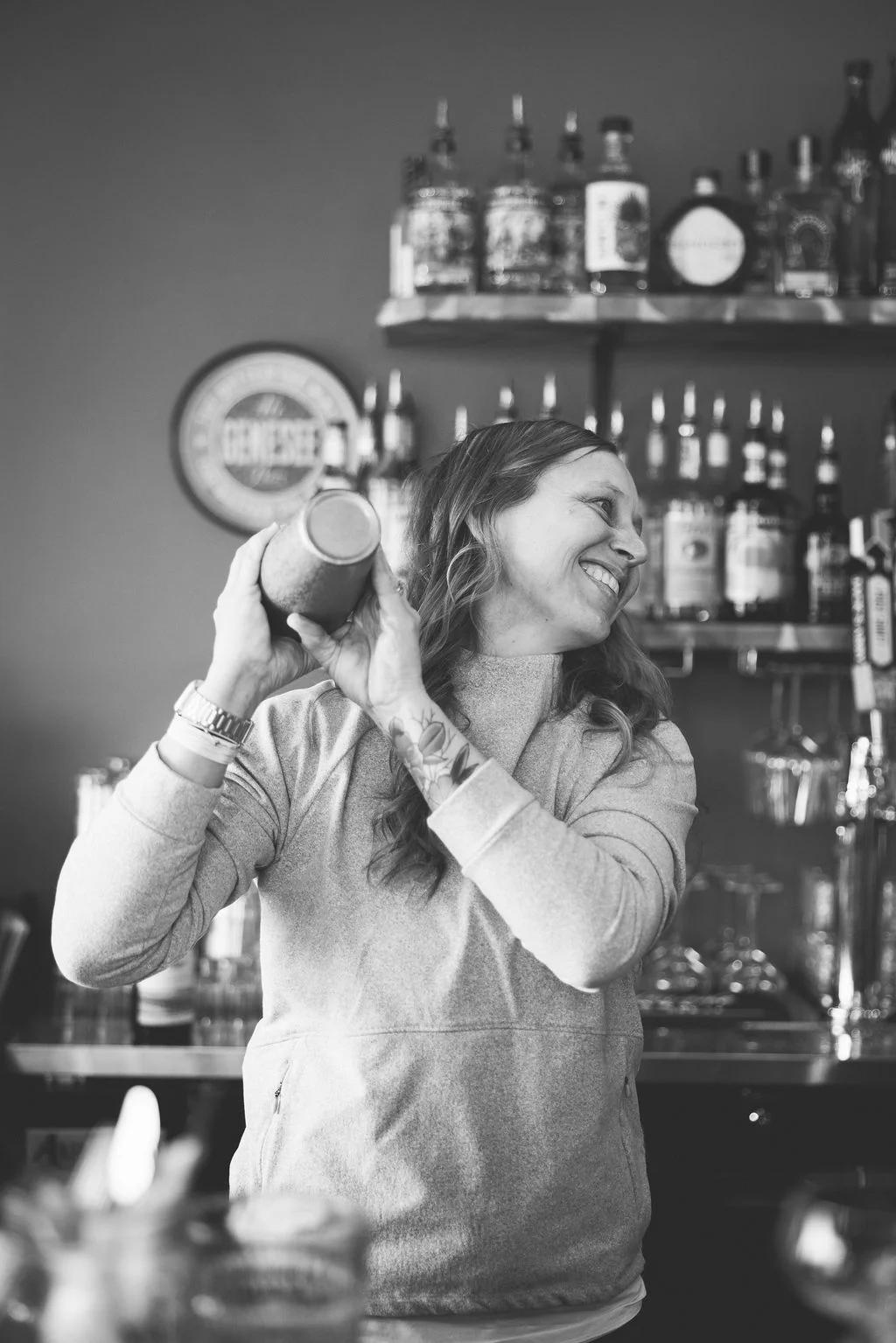 A bartender joyfully shaking a cocktail shaker, surrounded by bottles of spirits on a shelf. The image conveys a lively bar atmosphere.