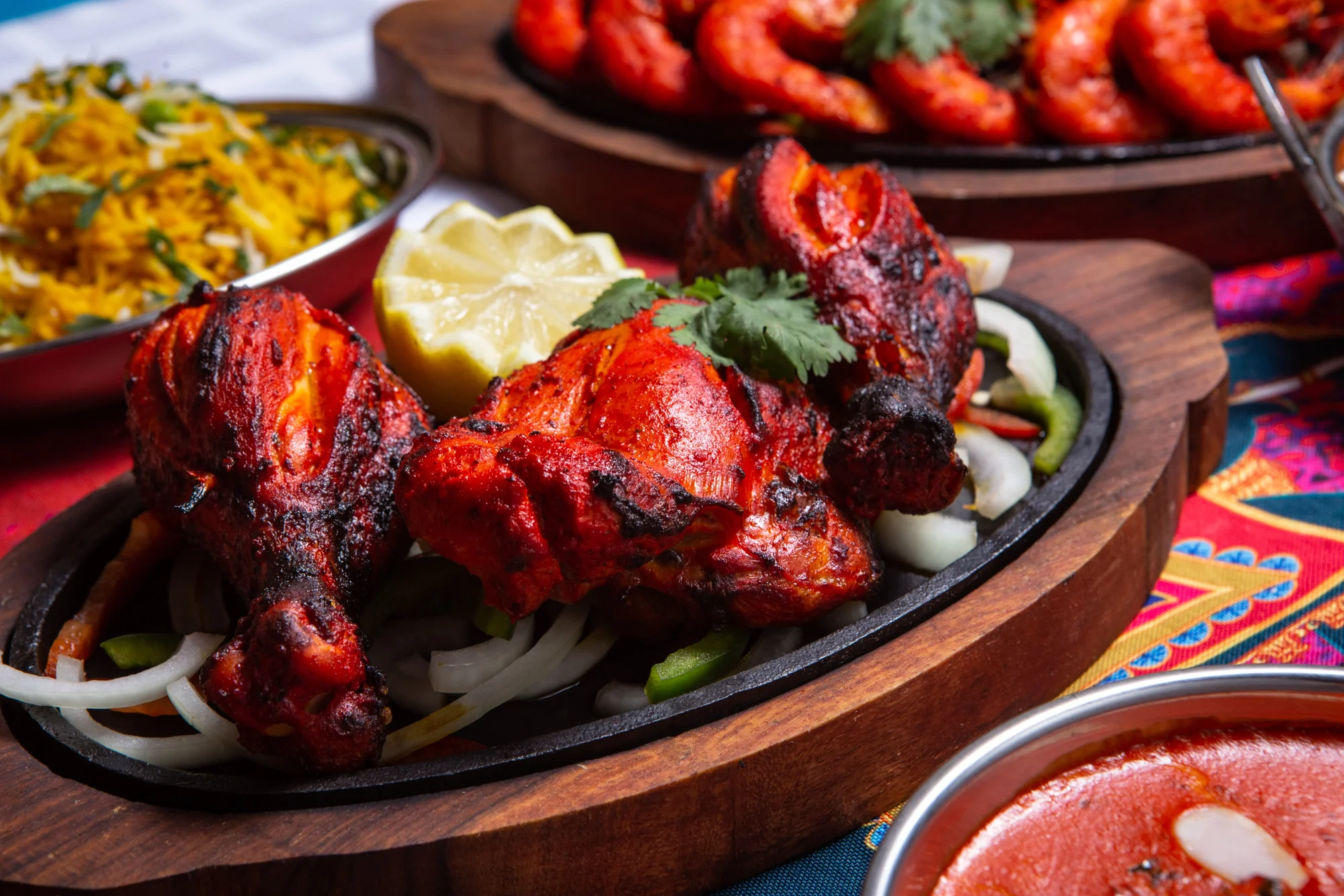 A vibrant dish featuring grilled chicken pieces, garnished with coriander and accompanied by lemon. The chicken is placed on a bed of onions and peppers, served on a sizzling platter.