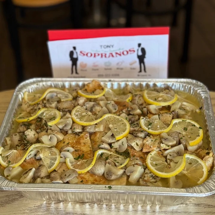 A dish featuring grilled fish topped with lemon slices and mushrooms, served in a metallic tray. The dish is accompanied by a restaurant sign in the background.