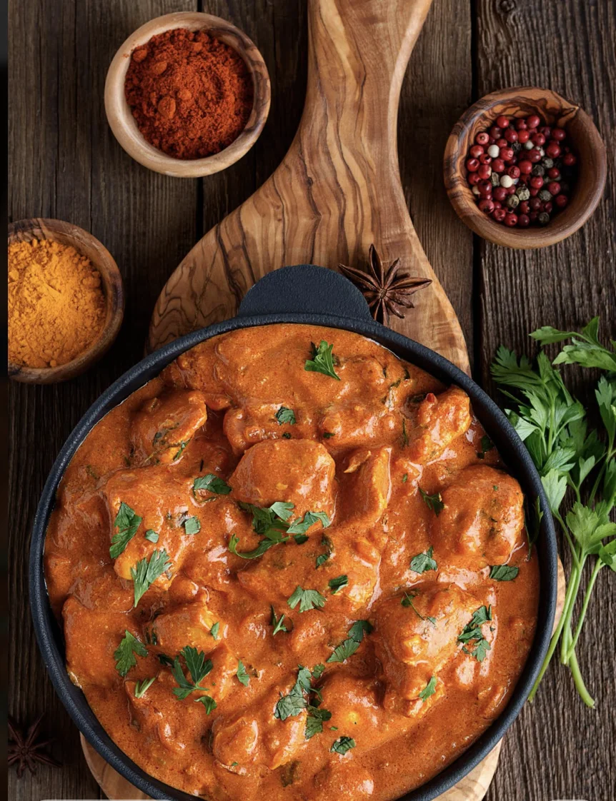 A rich and flavorful curry with chicken pieces, garnished with fresh herbs, served in a black pot along with spices in wooden bowls.
