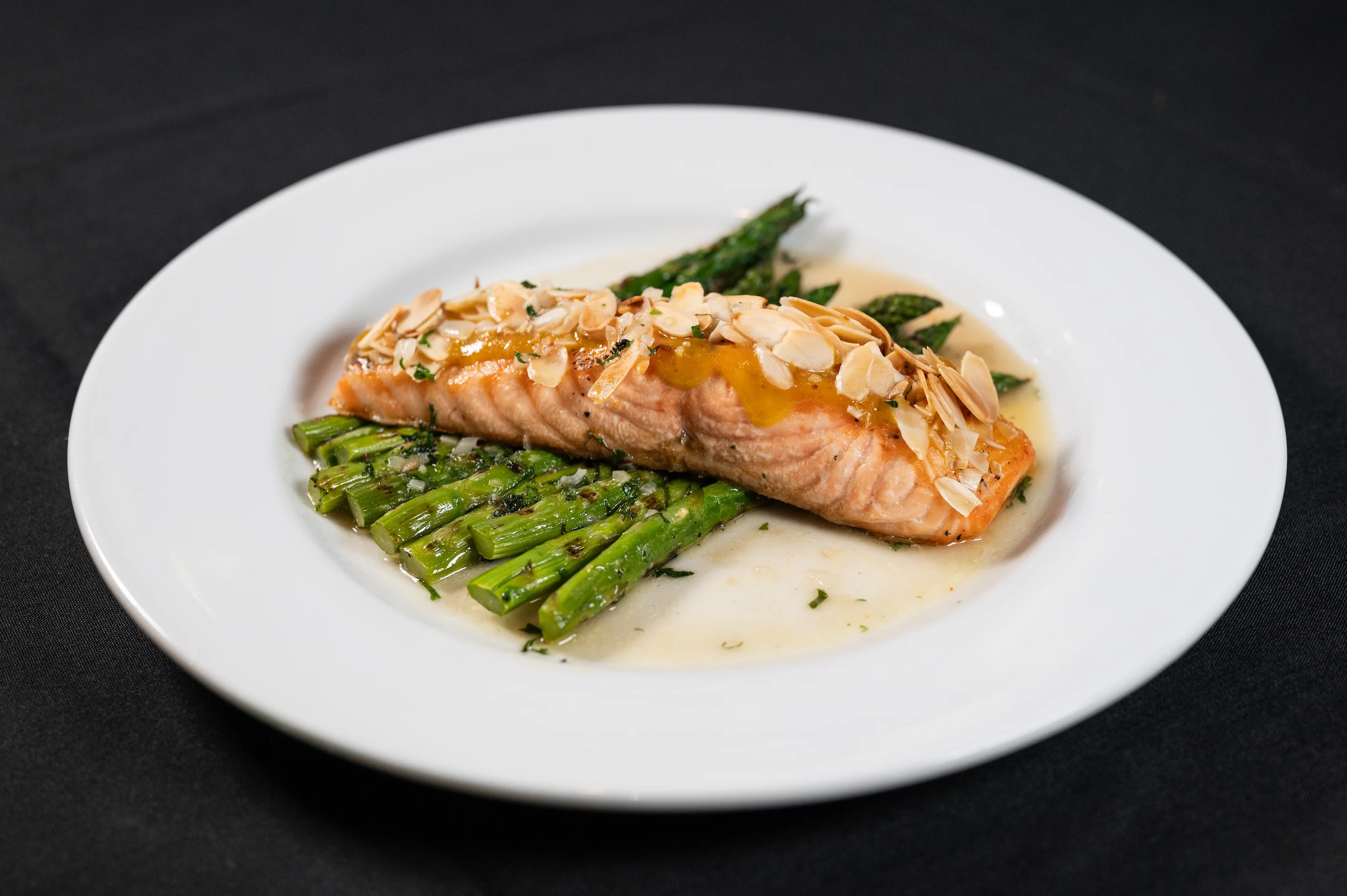 Delicious baked salmon fillet topped with almonds, served on a bed of sautéed asparagus, garnished with herbs and sauce served at Toscana on Main