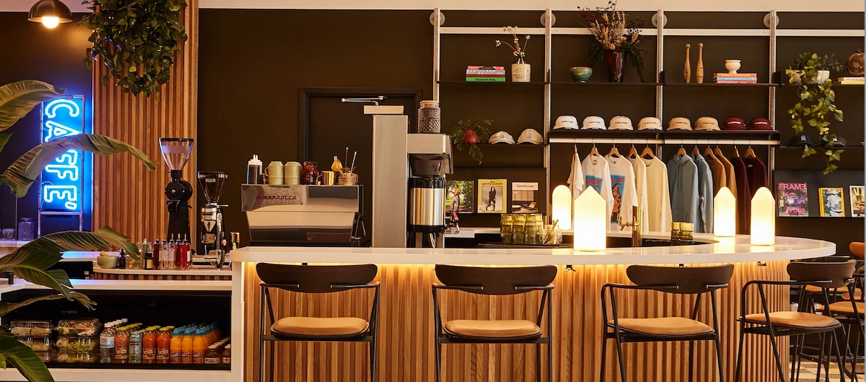 A modern café interior with a circular bar, shelves displaying merchandise, and a neon sign. The space is adorned with plants, creating a welcoming atmosphere.