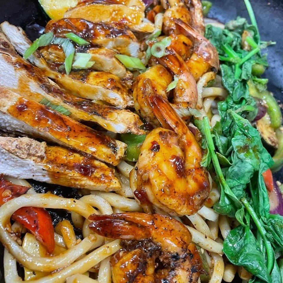 A colorful dish featuring grilled shrimp and chicken served over noodles with mixed vegetables.
