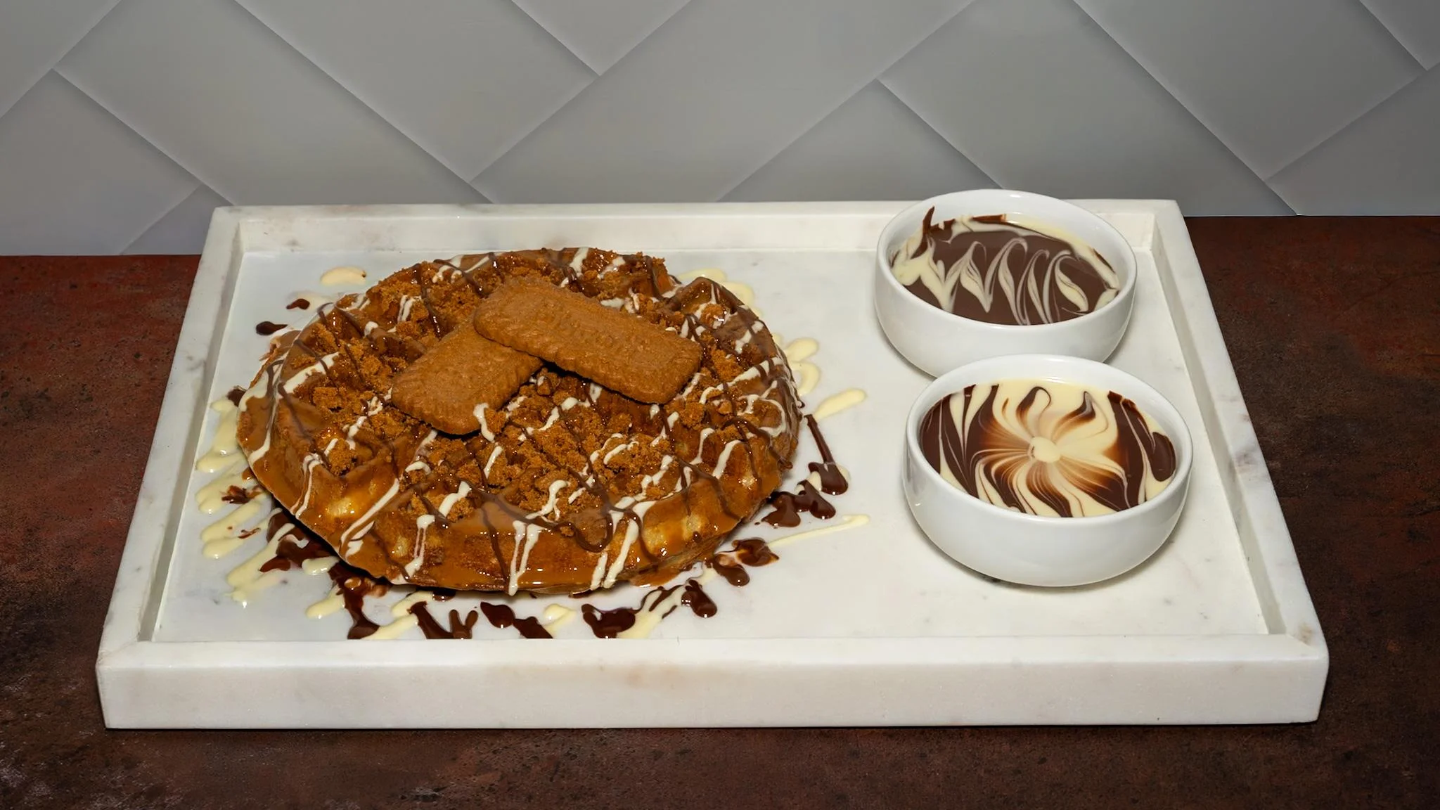 A dessert consisting of a waffle topped with cookie crumbs and drizzled with sauces, served with bowls of chocolate and creamy sauce.