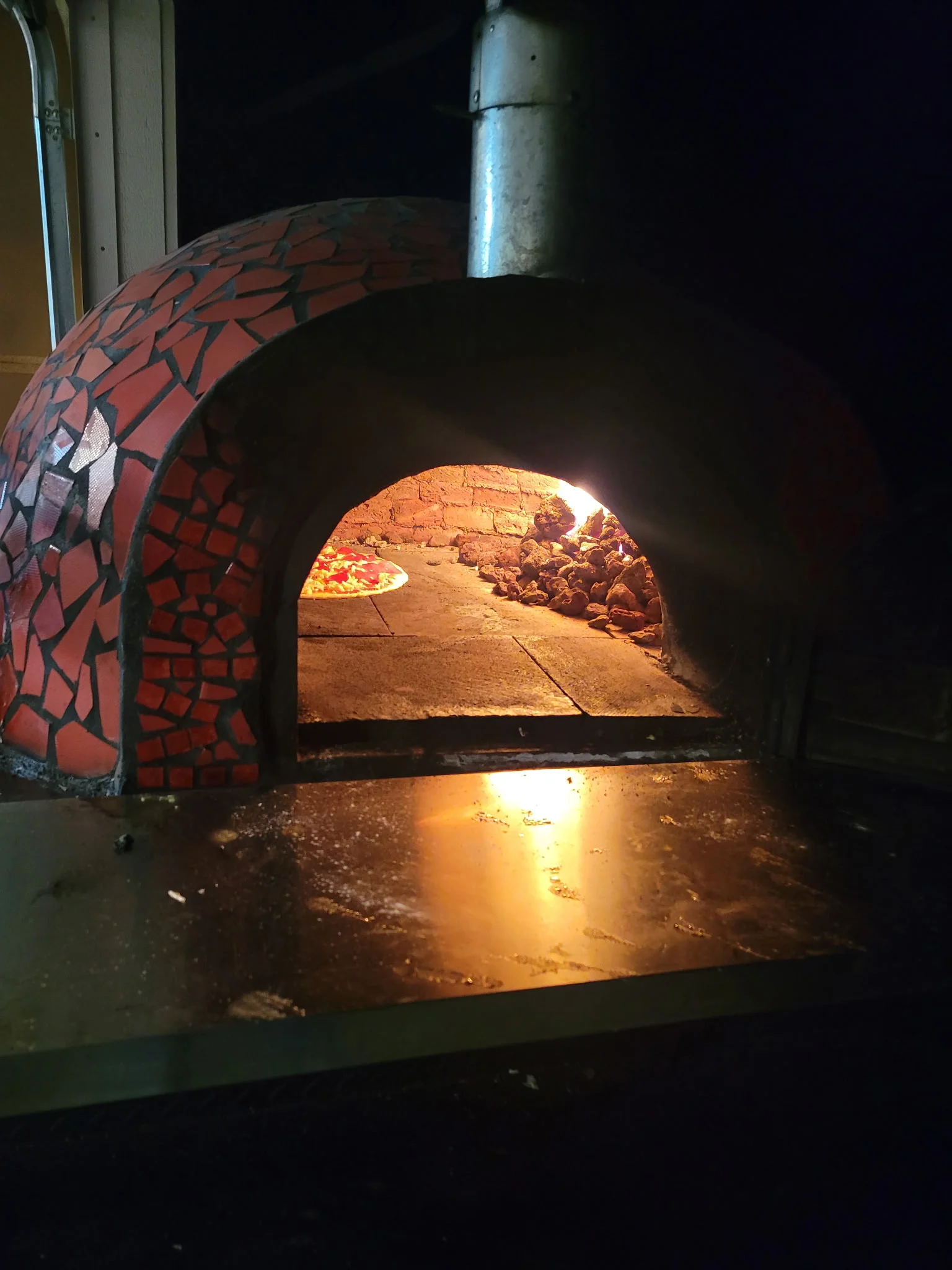 A person is working at a wood-fired oven, smiling as they prepare food. The oven features a colorful tiled design.