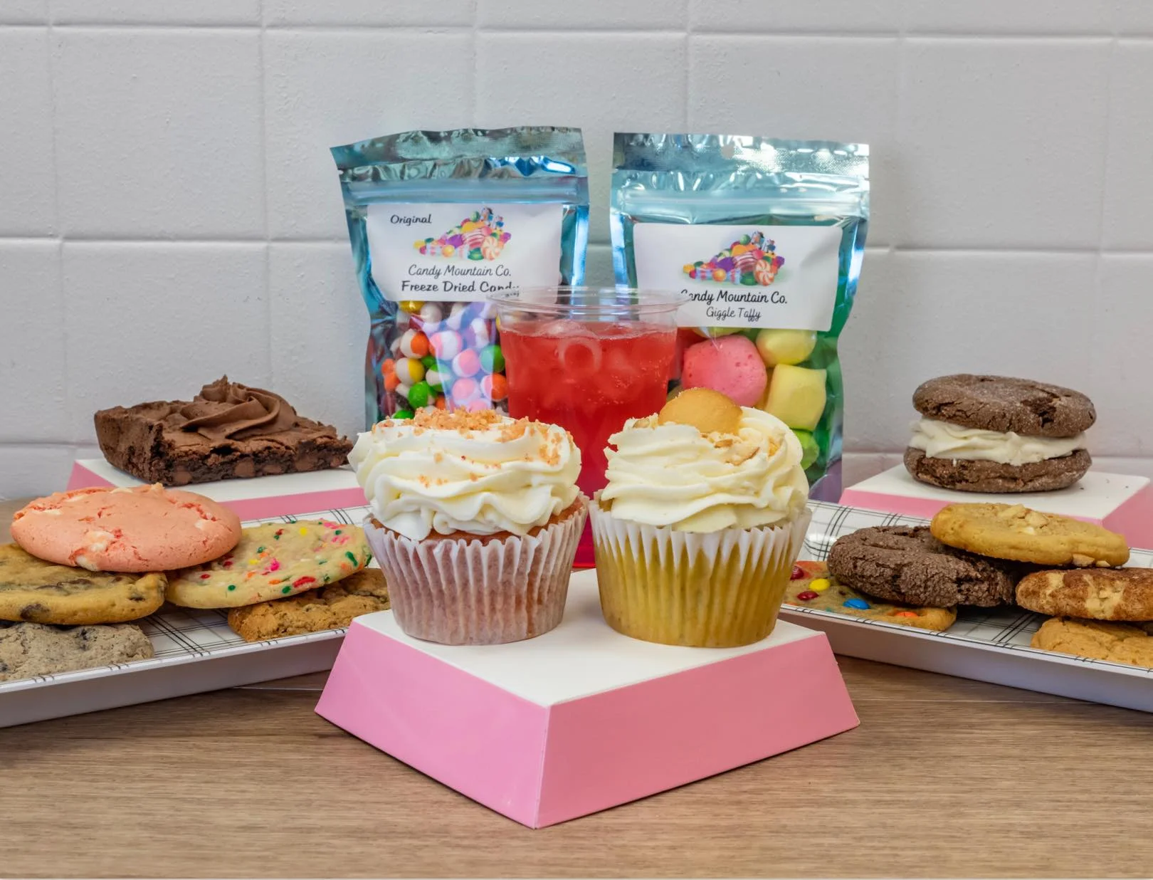 A dessert spread with cupcakes, cookies, brownies, and packaged candy, displayed with drinks on a table.