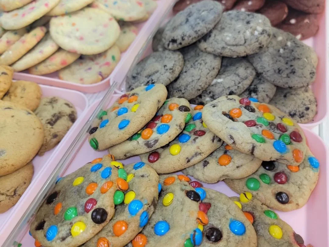 Trays of assorted cookies, including cookies topped with colorful M&M candies, chocolate chip cookies, and Oreo-style cookies.